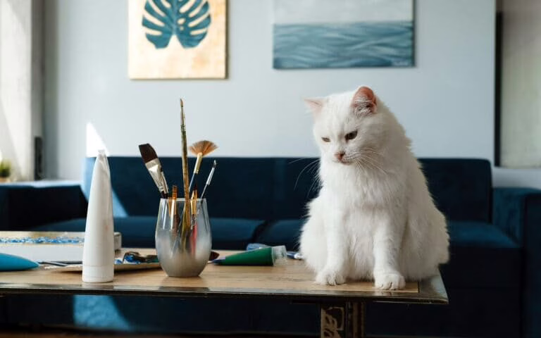 A cat on top of a table with paint brushes close to it,